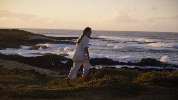 Um homem dá um passo em um penhasco gramado perto da costa, admirando as ondas do oceano quebrando contra as rochas escuras enquanto o pôr do sol dourado brilha ao longo do horizonte.