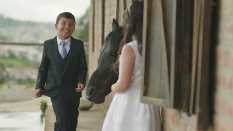 a boy in an elegant suit and a girl in a white dress tenderly caress a horse from a stable window.