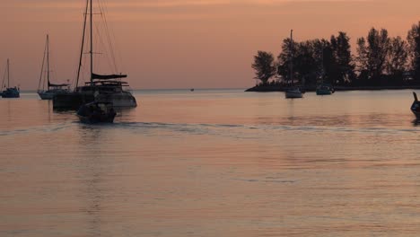 Beautiful-landscape-shot-capturing-shimmering-sunset-reflection-on-calm-water-surface-with-fishing-boat-cruising-forward-and-luxurious-yacht-anchored-in-the-bay,-langkawi-island,-malaysia