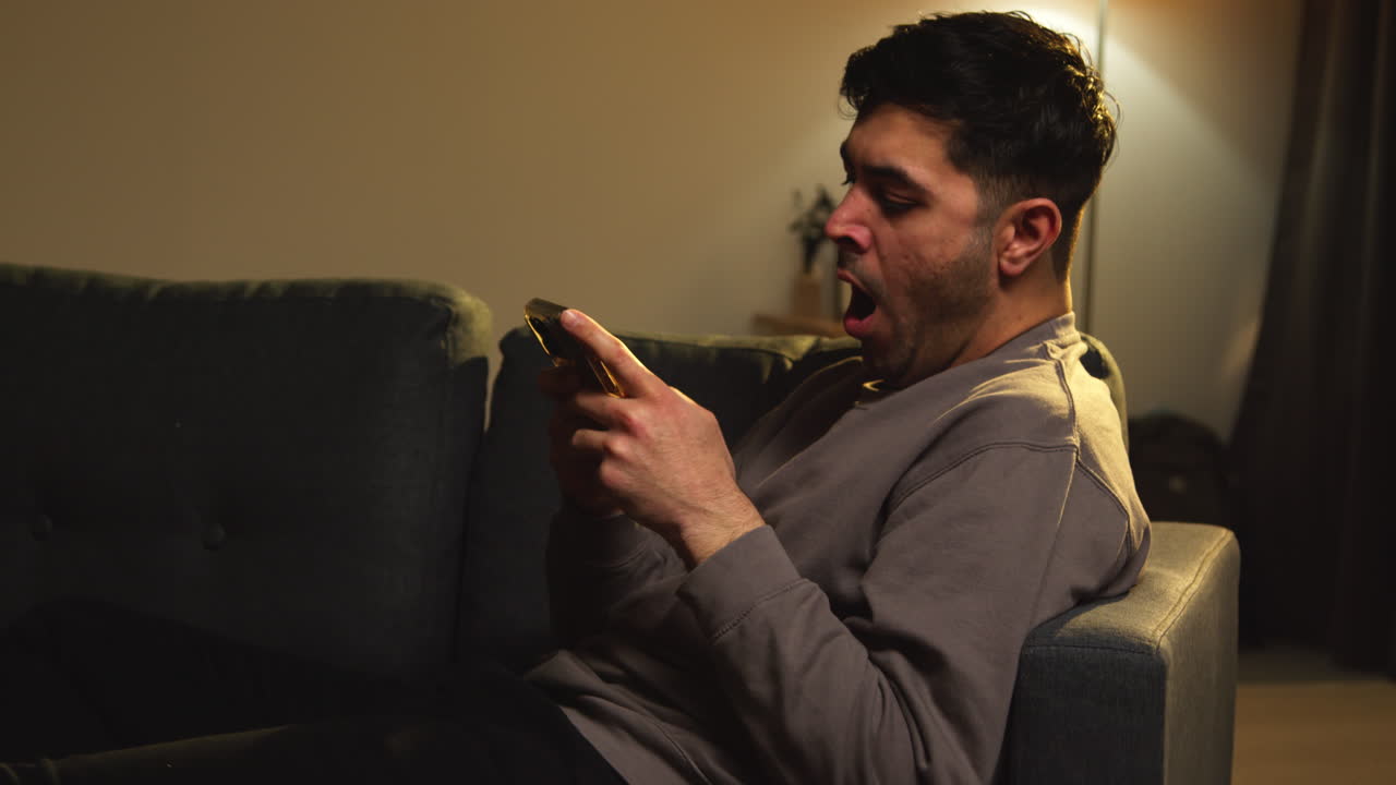 hombre cansado y bostezando pasando la noche en casa sentado en el sofá con el teléfono móvil desplazándose por internet o las redes sociales