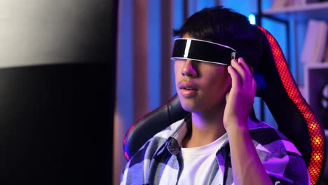 close up of asian boy streamer playing game, touching, and swiping experience virtual reality in futuristic goggles. personal computer on desk illuminated by rgb led strip light