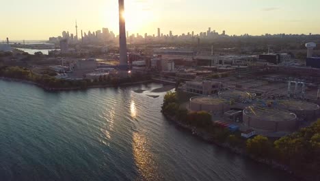 Aerial-Sunset-Wide-Shot-Sliding-Left-Over-Lake-Shoreline-Orbiting-Huge-Smokestack-Tower-That-Eclipses-The-Sun-With-Industrial-Factory-And-Downtown-Skyline-In-Background-In-Toronto-Ontario-Canada