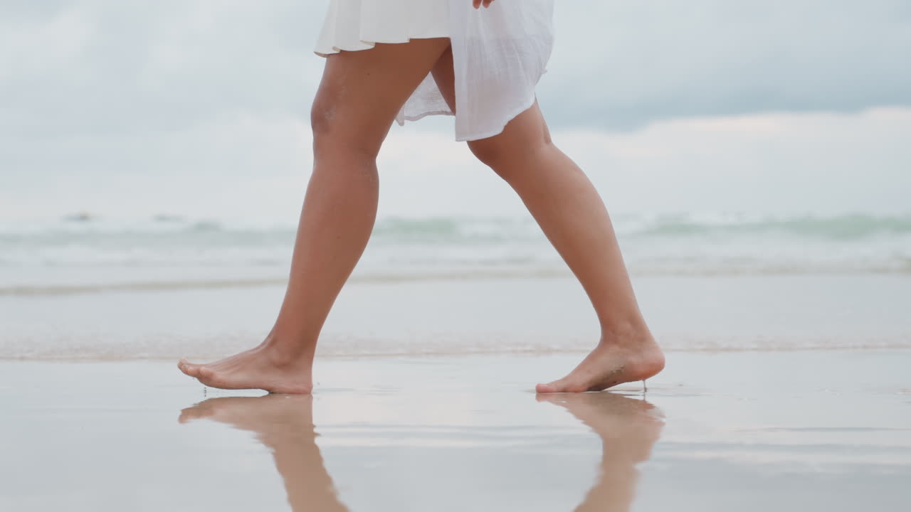 Passeggiata serena sulla spiaggia. Donna elegante sulla costa sabbiosa. Figura femminile calma e posata che cammina tra le onde dell'oceano. Donna aggraziata con movimenti fluidi riflessi nella sabbia bagnata lungo il litorale tranquillo