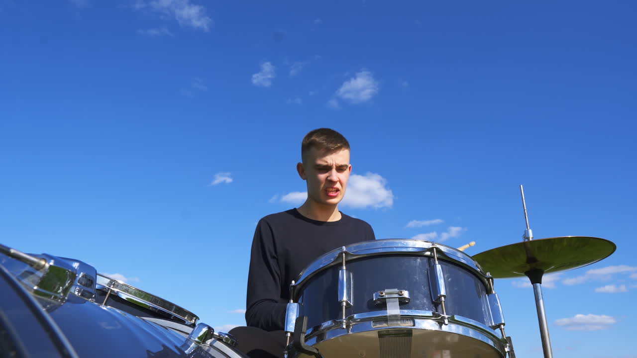 Young Caucasian man playing drums savagely. Emotional male musician hitting drums and cymbals with wooden sticks at blue sky backdrop. Low angle view.