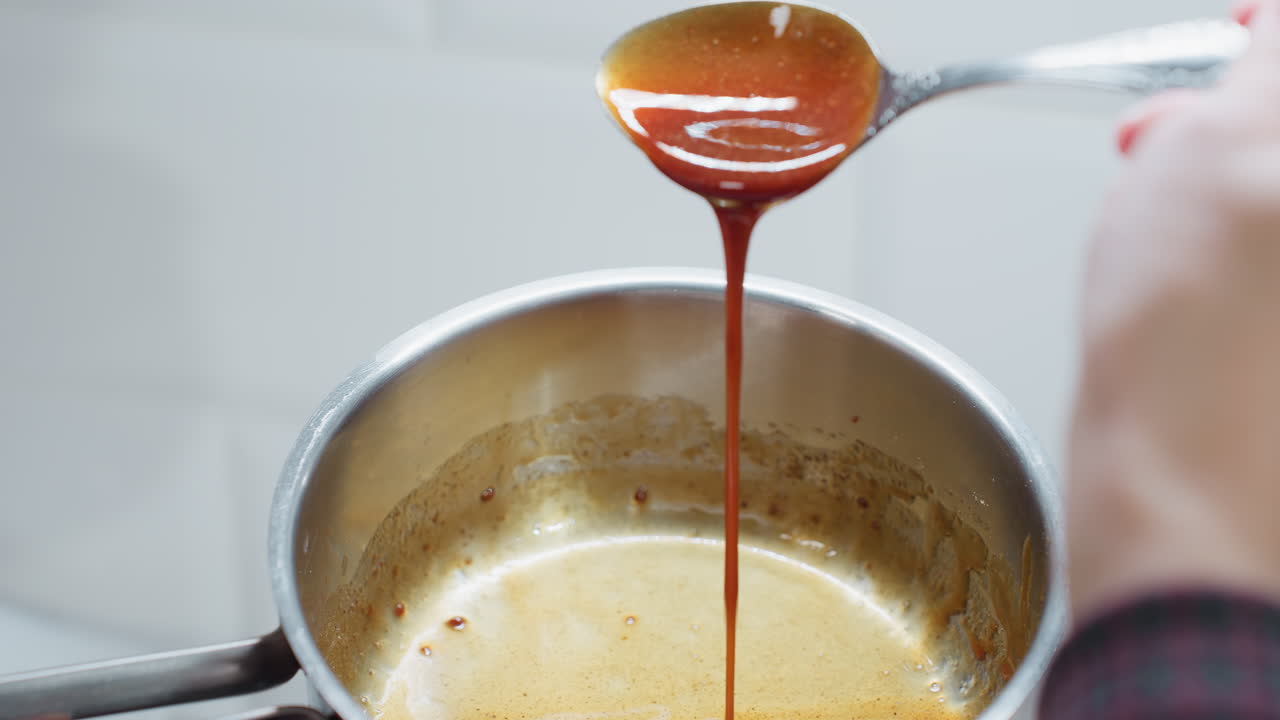 Close-up of person scooping and pouring rich red palm oil into a stainless steel pot, the oil flows smoothly, creating a glossy texture inside the pot, reflecting warm kitchen lighting