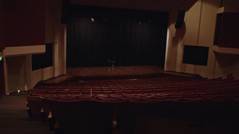 wide shot showing rows of empty red cushioned seats in a dark theater facing a wooden stage with black curtains and a stool