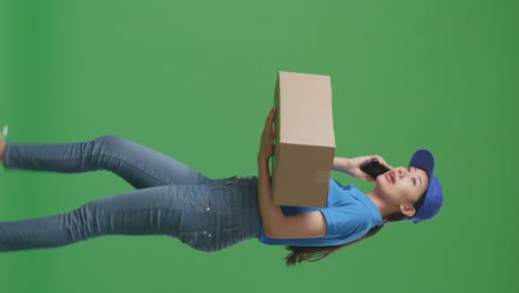 side view of asian female courier in blue uniform walking and talking on smartphone while delivering a carton on green screen background in the studio