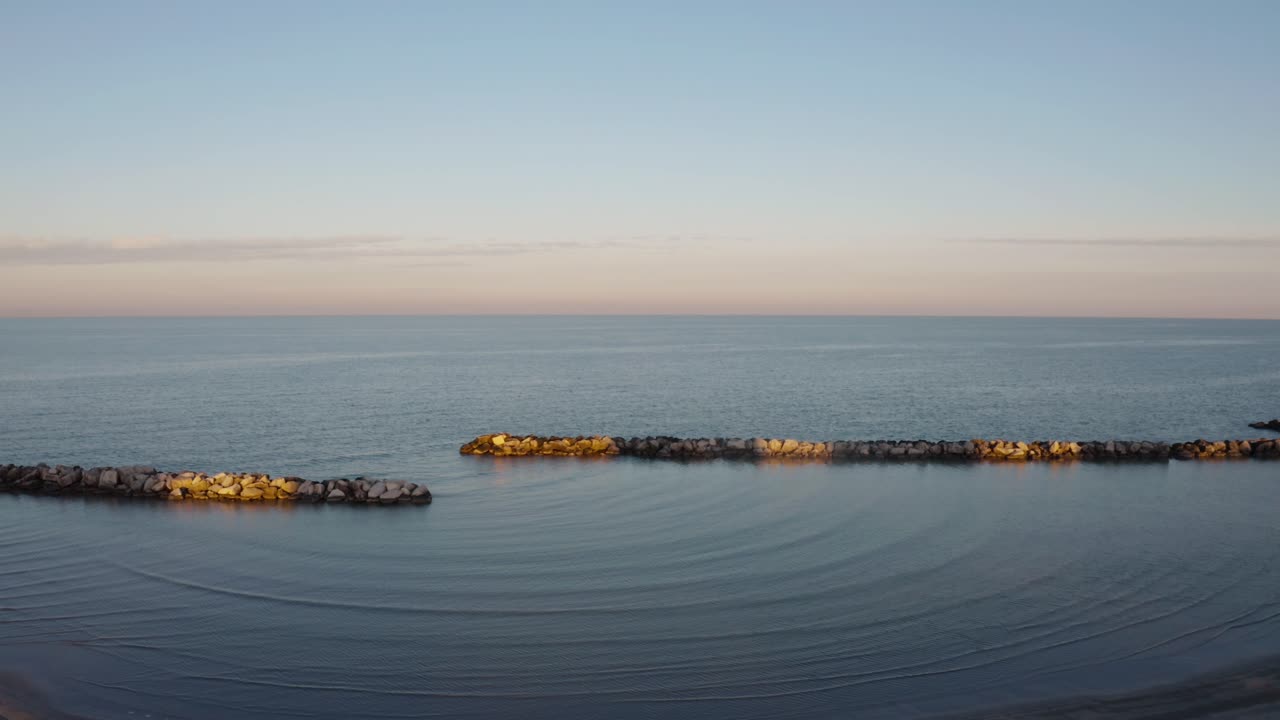 Ampia vista costiera di Rimini con mare calmo, spiaggia sabbiosa, figure distanti e frangiflutti illuminati dal sole sotto un cielo serale dai colori pastello tenui.