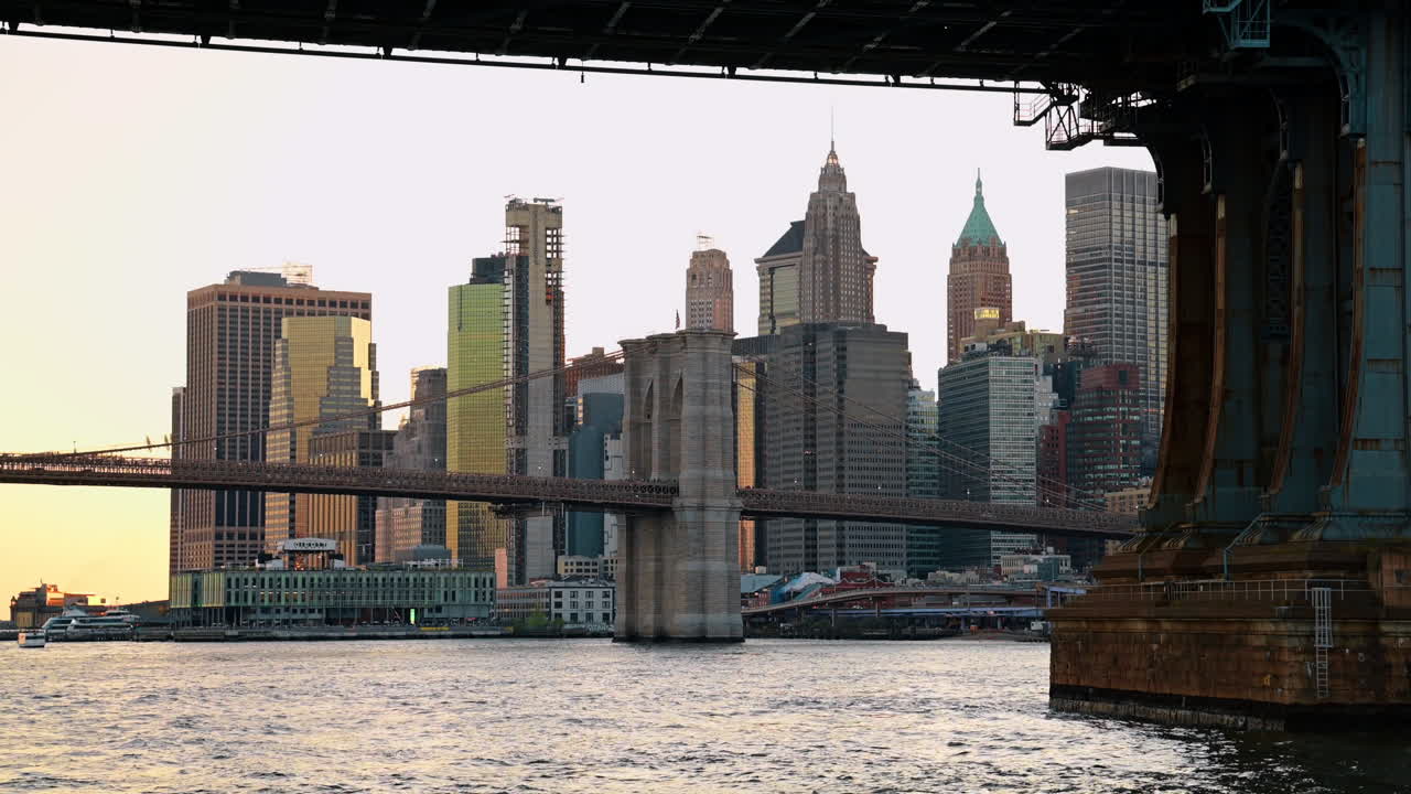 El horizonte del Bajo Manhattan y el Puente de Brooklyn vistos bajo el Puente de Manhattan. Rascacielos del Bajo Manhattan y el Puente de Brooklyn enmarcados por la estructura de acero del Puente de Manhattan al atardecer.