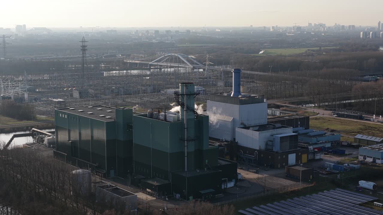 Industrial power station with a tall chimney releasing smoke into the atmosphere, a large solar panel farm nearby, and a hazy city skyline visible in the distant background