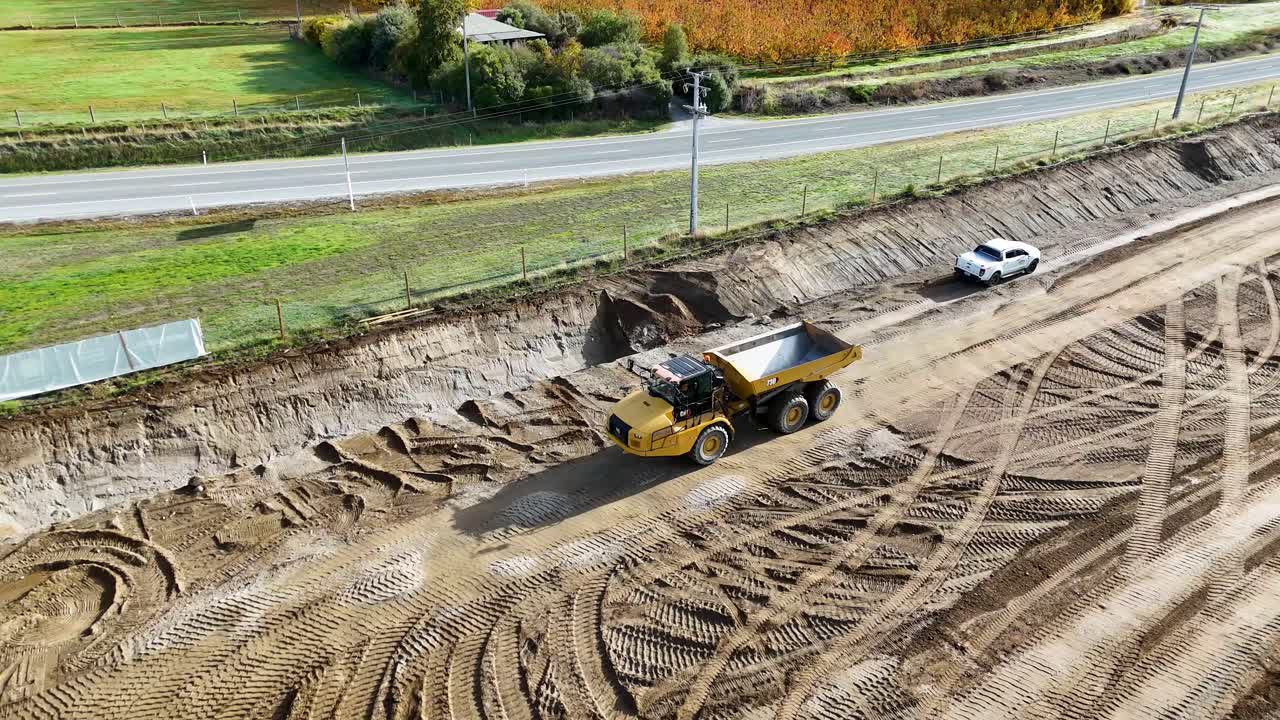 Drone footage captures an excavator and dump truck at a construction site in Cromwell, New Zealand, under bright daylight