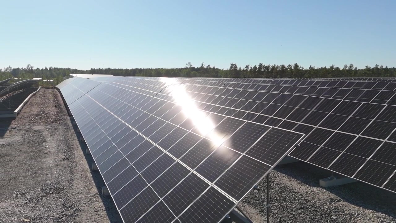 Panorâmica aérea em movimento para a esquerda ao longo de uma fileira de painéis solares bifaciais refletindo a luz solar em uma Fazenda Solar na Finlândia, raio de sol