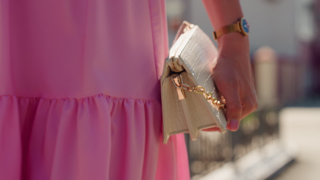 Abito rosa, clutch in primo piano in mano, borsetta beige con catena e chiusura dorate che catturano la luce del sole, orologio da polso scintillante, recinzione e sfondo cittadino bokeh, dettaglio di accessori estivi editoriali e strada elegante