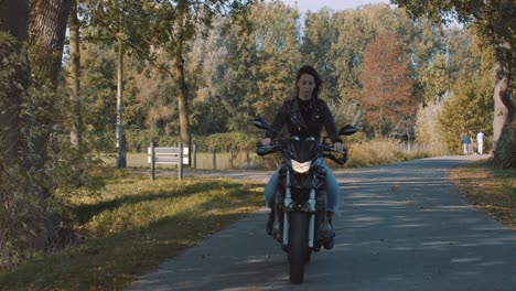 mujer joven europea muy sonriente conduciendo una moto con chaqueta de cuero en el bosque con hojas de otoño doradas vibrantes y coloridas en el día soleado-1