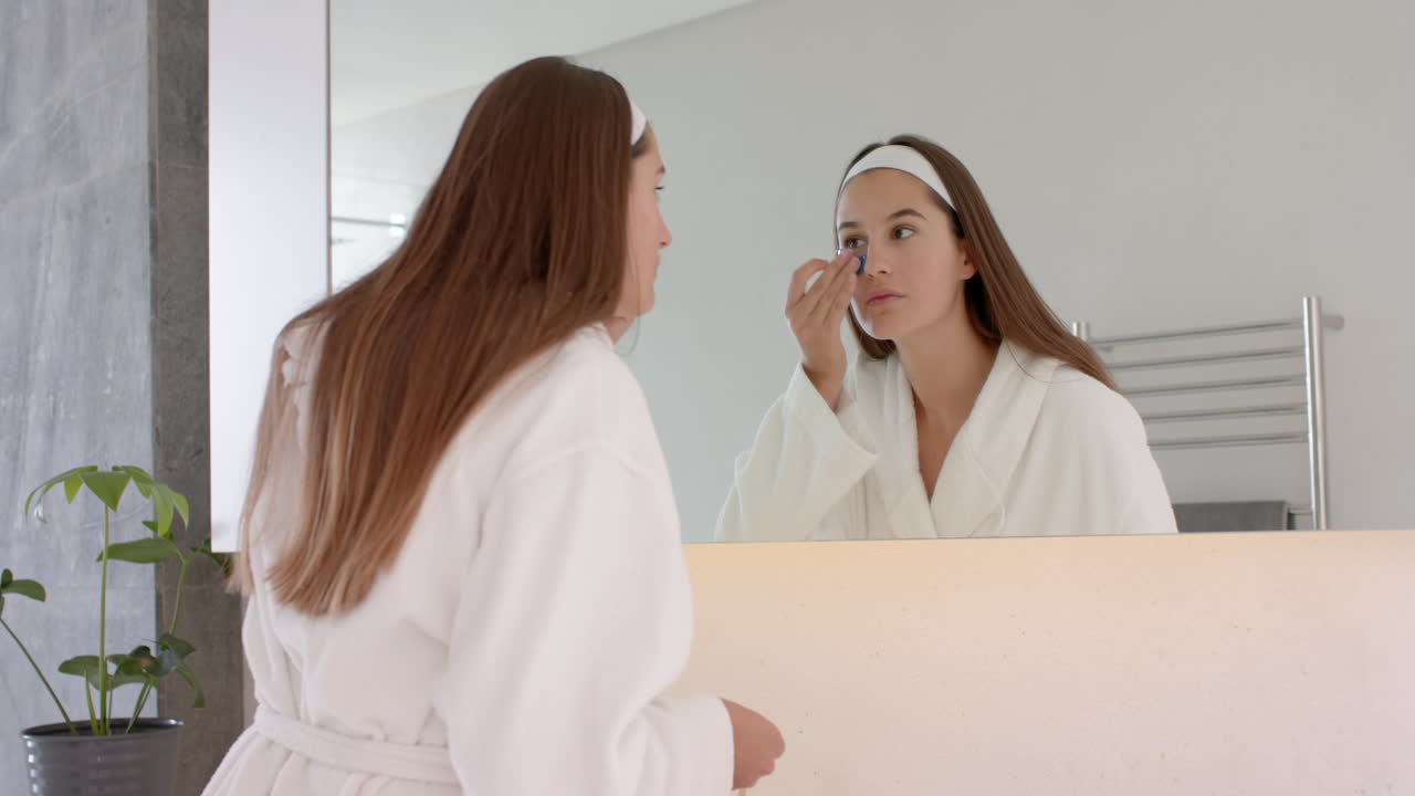 Applying skincare product, woman in bathrobe looking in mirror at home spa