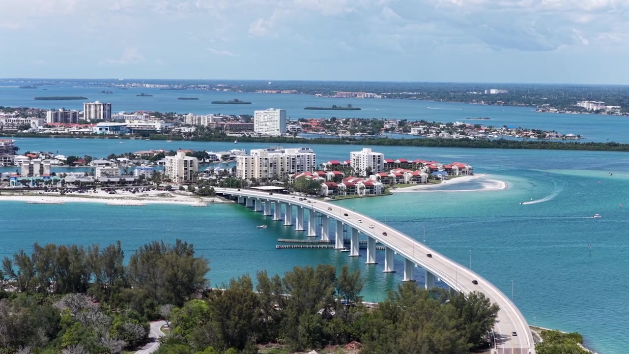 Ponte curvo attraversa acque turchesi collegando isole barriera, condomini con tetti rossi e spiagge sabbiose che fiancheggiano il traffico verso uno skyline lontano. Clearwater Beach, Florida, USA. Ampia ripresa aerea con drone