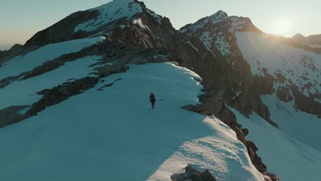 Drone-shot-from-behind-of-a-female-mountaineer-hiking-across-a-scenic-snowfield-at-sunrise-in-the-mountains-of-South-Tyrol,-Italy