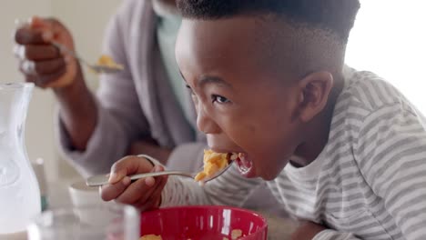 happy african american grandfather having breakfast with grandson in kitchen, slow motion