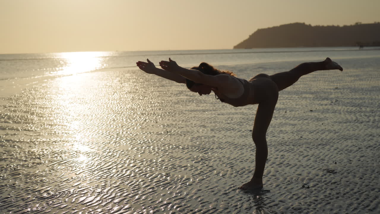 Kvinde dyrker yoga i balanceposition på stranden ved solnedgang