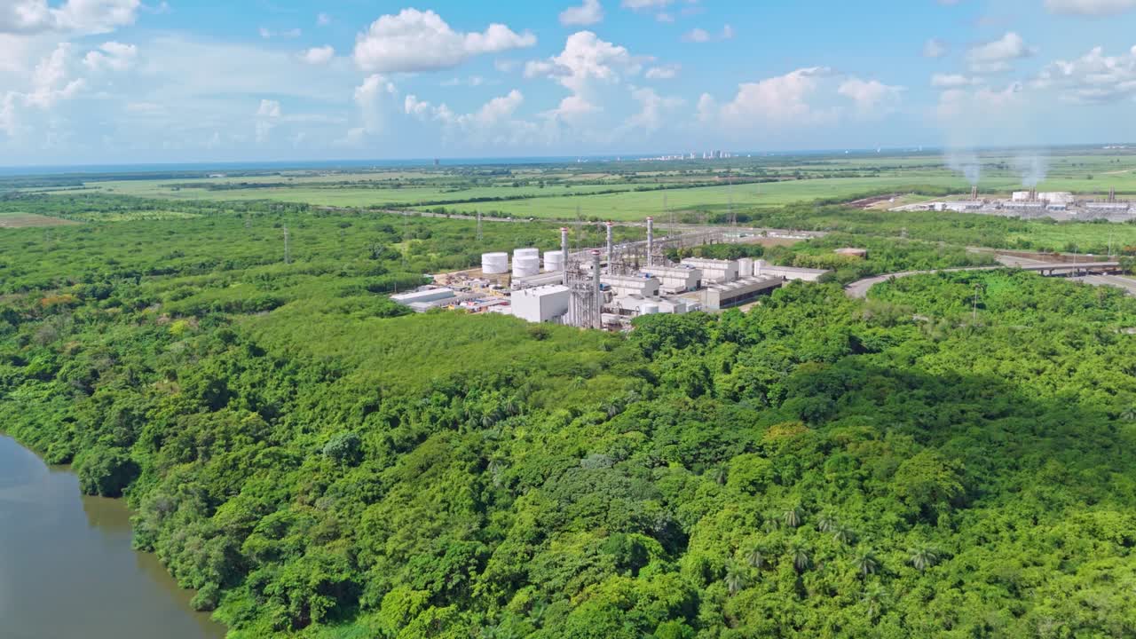 Energa 4 power plant and storage tanks surrounded by lush green forest and river, contrast between industry and nature, Dominican Republic. Aerial forward