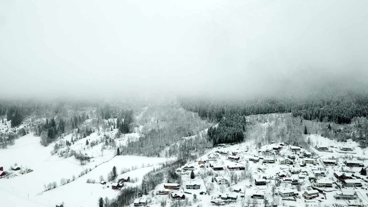 Mystic aerial flight towards dense fog and snowy mountains in backdrop. Snow-covered buildings in valley during cold winter day in Norway.