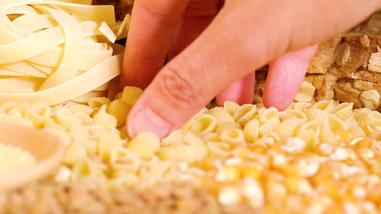 A hand picks conchiglie pasta from a grain assortment, set against a backdrop of bread and pasta, under warm lighting