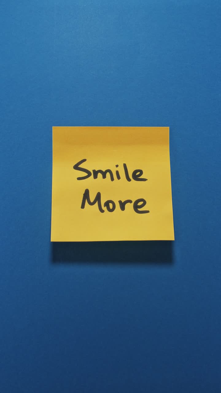 Mani che attaccano un post-it "Sorridi di più" su una parete blu, evidenziando una mentalità positiva.