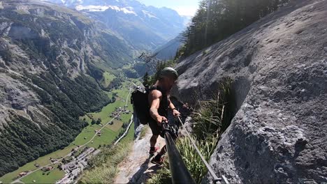 strong man walks very carefully on the small path on the edge of the cliff of via ferrata route with a steep and tall mountainside right behind him. beautiful and lush valley below in the alps.