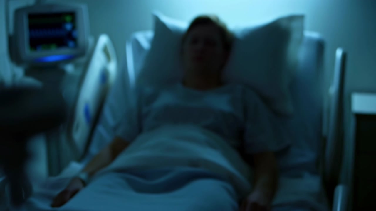 Primer plano de tubos médicos y una pinza junto a un interior de hospital difuminado con una persona indistinta bajo una luz azul fría.