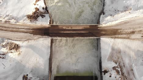 bird's eye view of small dam icy floodgate, flowing through cold white stream - aerial shot