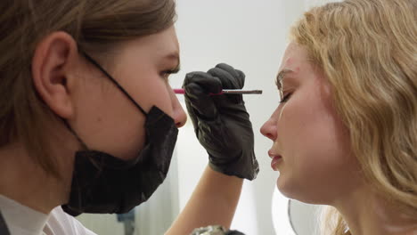 beautician wearing black gloves carefully applies cream to client eyebrow using pink-handled brush while client remains relaxed in bright, softly lit room with minimal background details in view