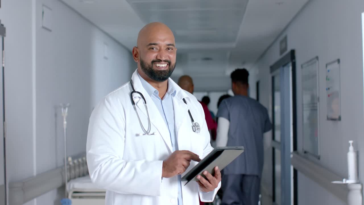 Ritratto di felice dottore di origine mista in camice da laboratorio, che usa un tablet nel corridoio, al rallentatore.