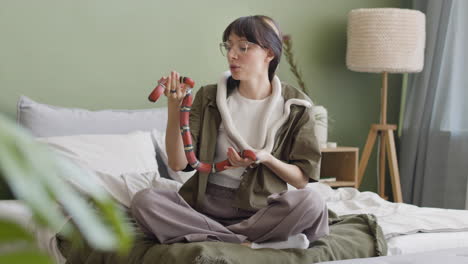 happy young woman holding her two pet snakes while sitting on bed at home