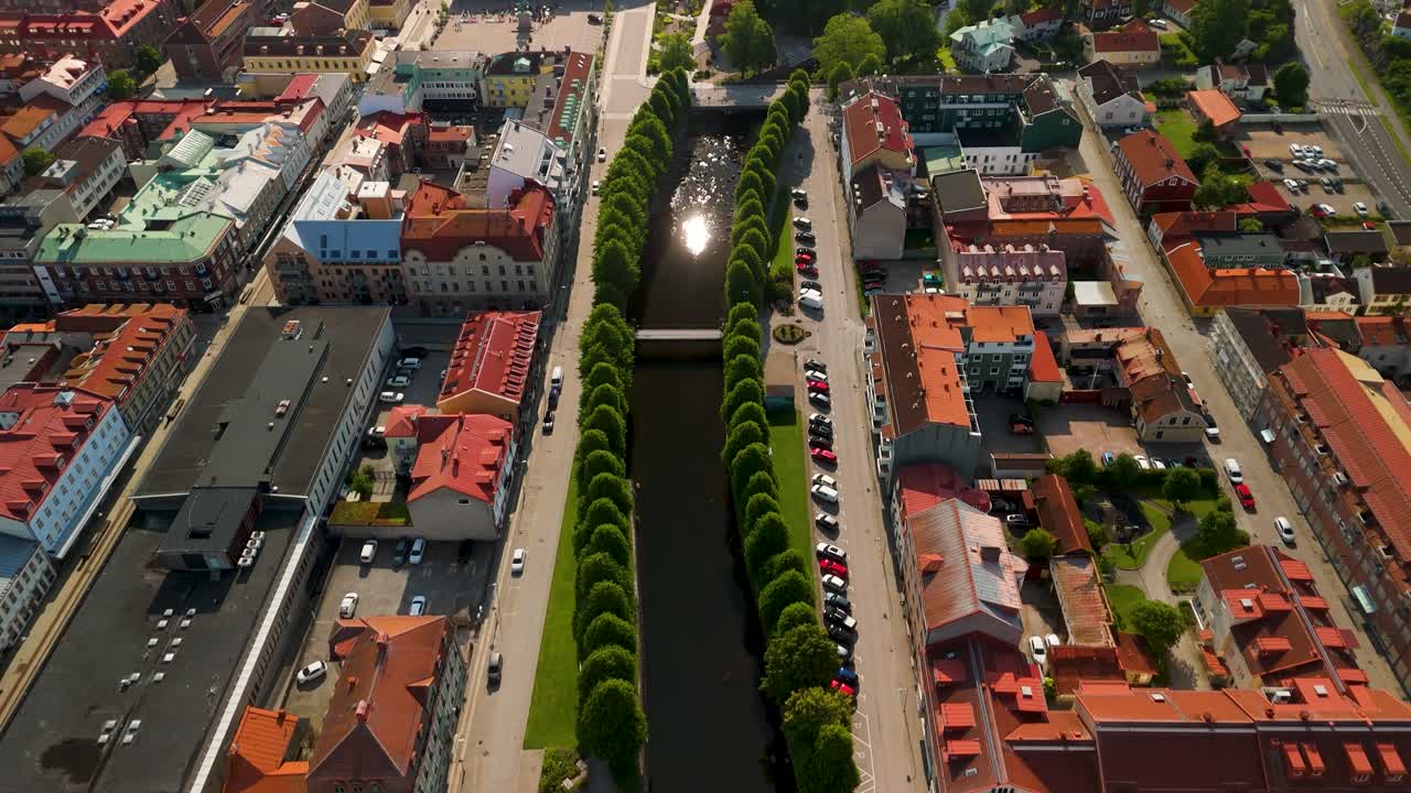 Drone che vola lungo il fiume Bäveån costeggiato da alberi verdi, inclinando la telecamera verso l'alto per rivelare il centro di Uddevalla e il campanile della chiesa sotto un cielo azzurro e limpido