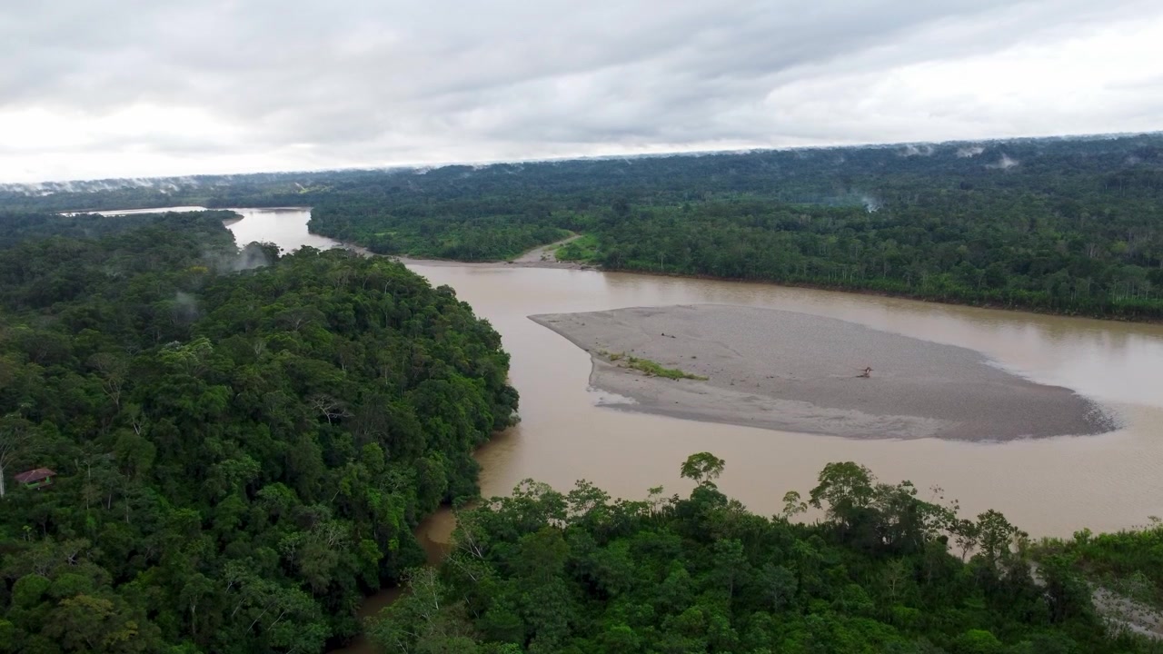 Aerial drone footage of a large sandbar in the middle of a wide brown Amazon river flanked by dense rainforest in Ecuador under a grey cloudy sky.