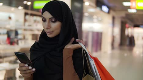 Attraktive-Muslimische-Frau-Mit-Schwarzem-Hijab-Kopftuch,-Die-Im-Einkaufszentrum-Spaziert-Und-Smartphone-Benutzt-1