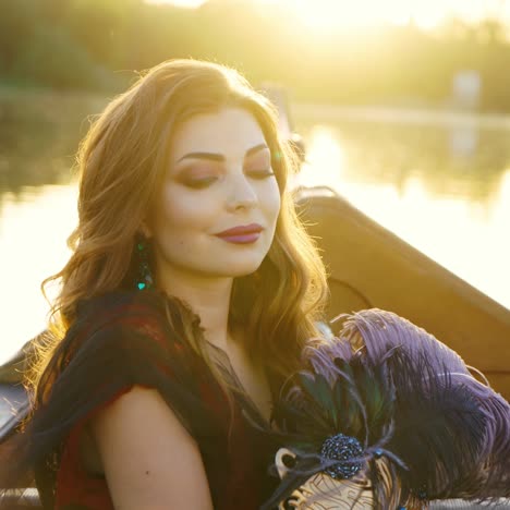 a pretty woman is holding a carnival mask which decorated with purple and black feathers. the girl tries on a mask in anticipation of a carnival in a gondola at sunset. blurred background