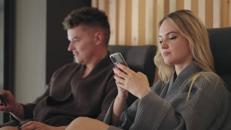 blonde-woman-and-her-husband-are-resting-together-in-spa-salon-relaxing-after-treatments-in-loungers-and-viewing-social-media