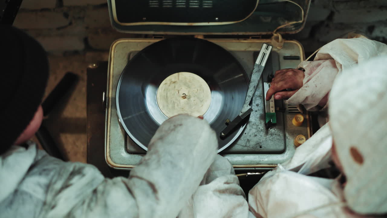 Overhead view of people in protective clothing adjusting vinyl record on old record player, one hand touching disc while another moves lever, symbolizing retro sound