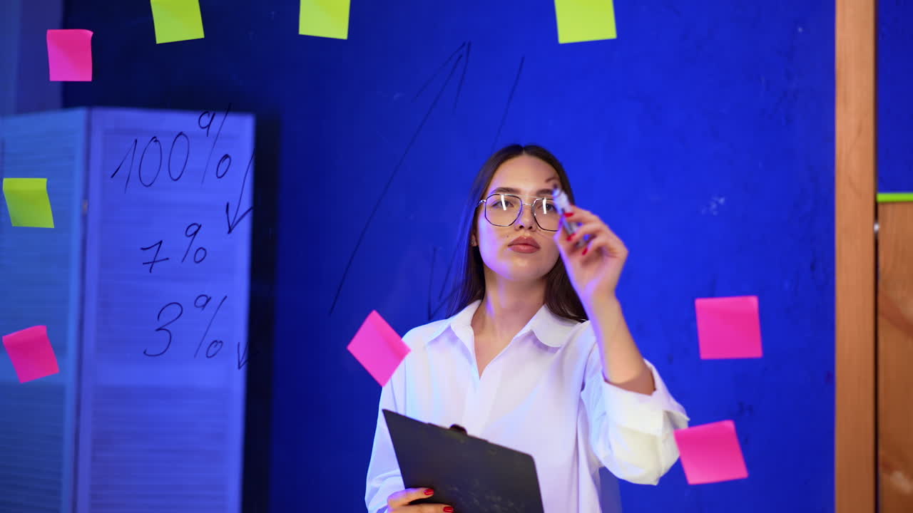 Concentrated business lady writing on the glass wall with marker. Woman writes numbers from the clip board.