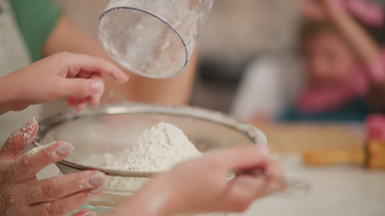 close up of person finishing flour pouring into sieve while child reaches hand inside glass jar to remove stuck flour as adult gives final shake, with playful kitchen activity in soft background blur