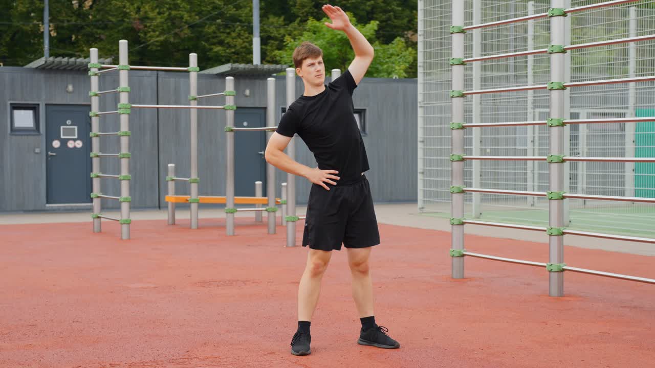 Young man stretching body in outdoor calisthenics park, performing warm up exercise for side bend and flexibility, preparing for fitness workout, static camera, slow motion