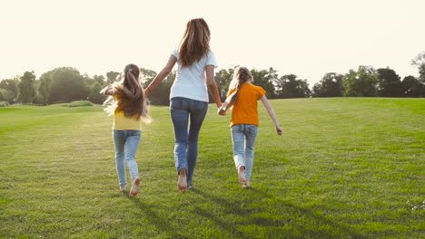 back view of mother and her two little daughters holding hands and running on green grass field in the park 1