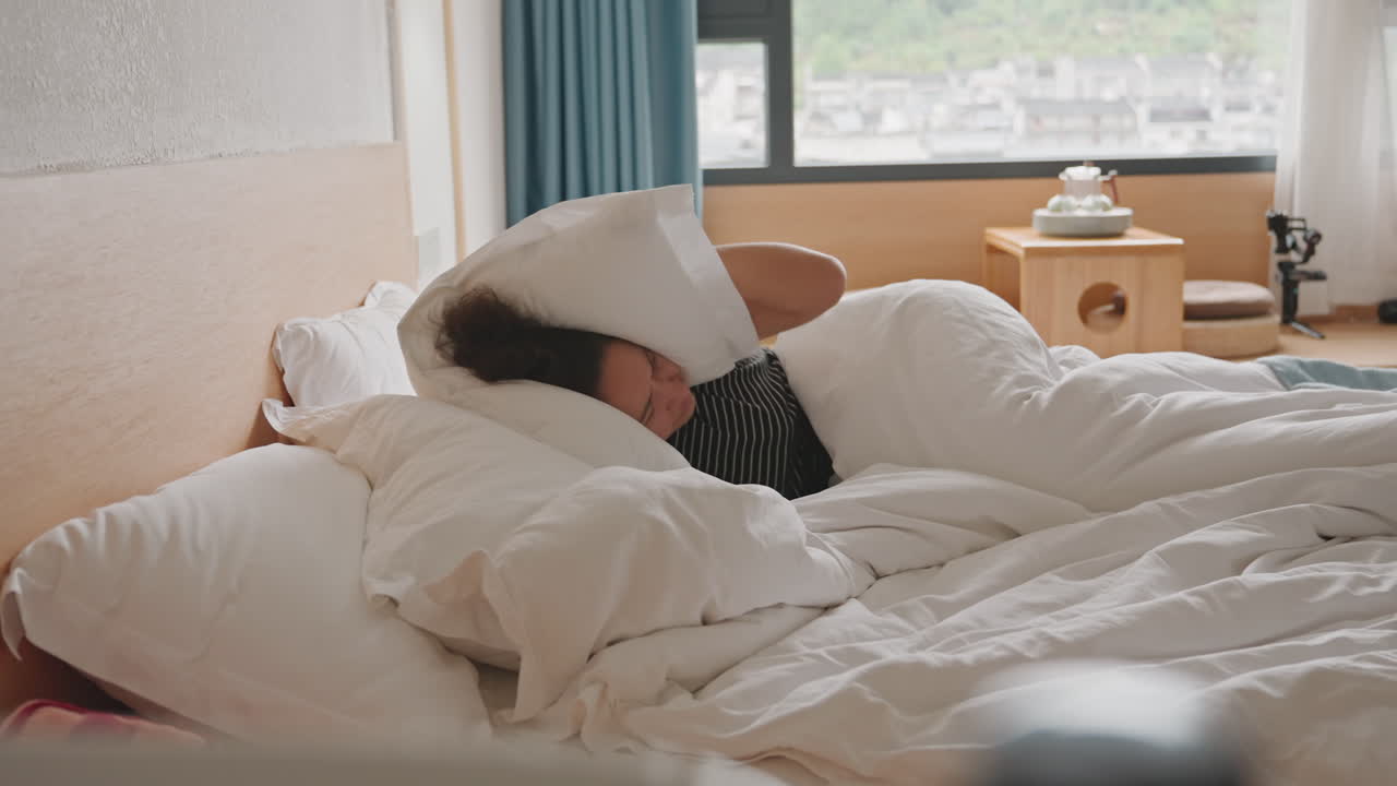 Overvældet hotelgæst gemmer sig under pude på rodet seng, totalbillede viser kvinde, der trækker pude over ansigtet ved vinduet med byudsigt, sengelampe og rejsetaske antyder ophold, dagslys oversvømmer rummet