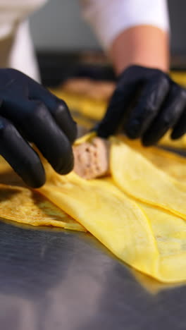 yellow thin crepe being rolled around meat stuffing. female gloved hands prepare semi-cooked dish for breakfast. vertical video