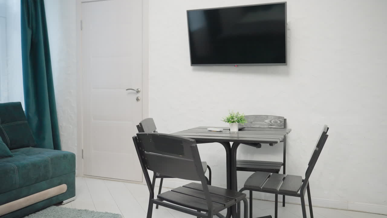 Cozy apartment with green velvet sofa, black dining table, small potted plant on the table, and flat-screen TV mounted on the wall. Light-filled space with large windows and teal curtains