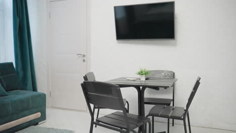 cozy apartment with green velvet sofa, black dining table, small potted plant on the table, and flat-screen tv mounted on the wall. light-filled space with large windows and teal curtains