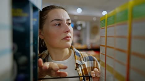 Close-up-of-a-happy-brunette-girl-in-a-plaid-shirt-looks-at-the-top-shelf-and-chooses-the-product-she-needs-during-her-shopping-in-the-supermarket