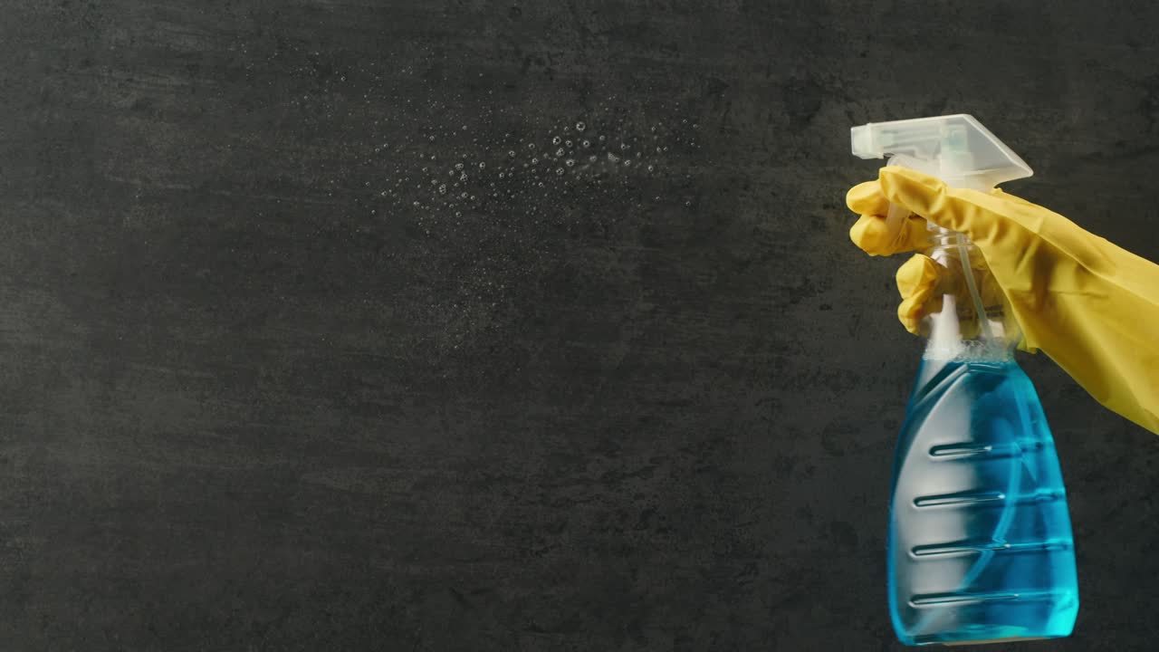 Mano con guanto giallo che spruzza un liquido detergente su una superficie scura