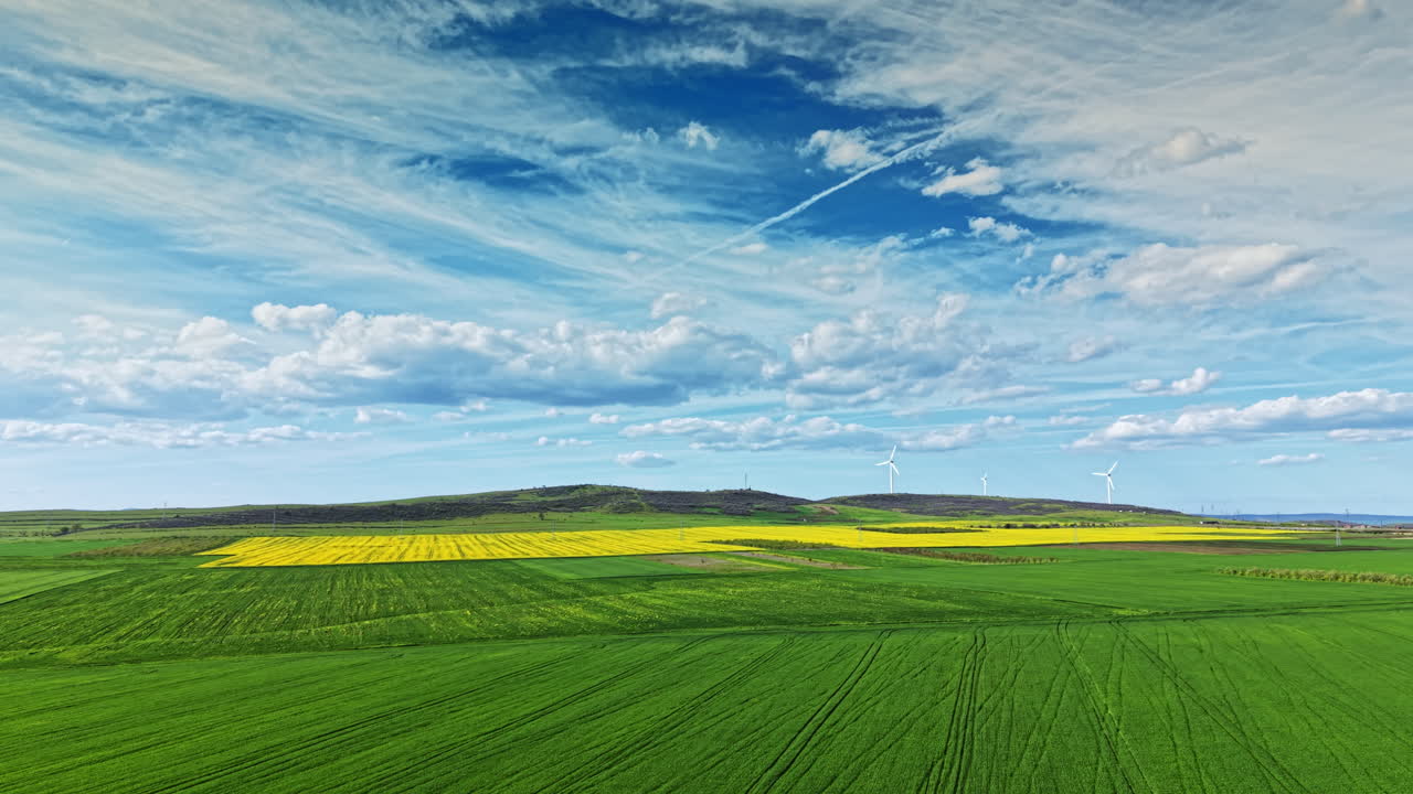 Fields of green and yellow stretch across the landscape under a bright blue sky with clouds and wind turbines scattered in the distance